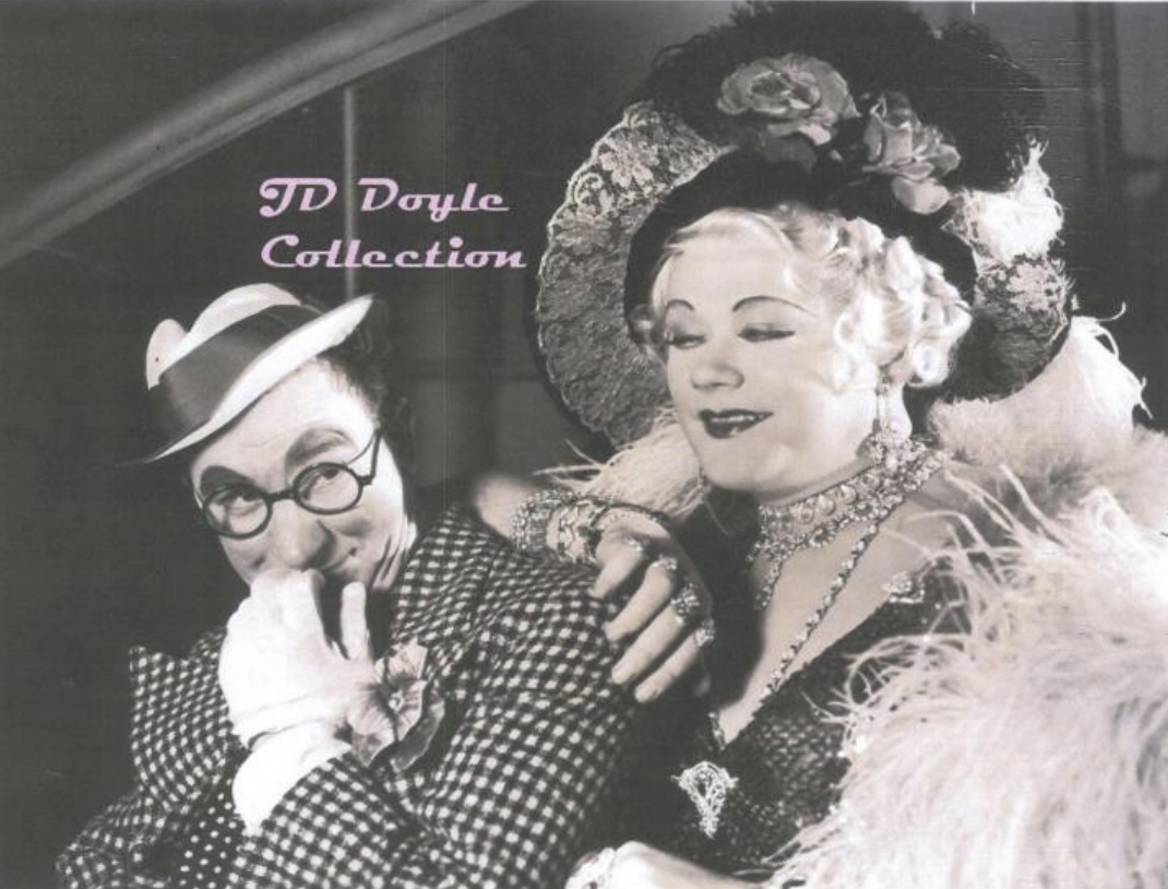 Black and white photograph from the movie "Arizona to Broadway" featuring a man in a hat laughing behind his hand while Gene Malin dressed in feather boa and hat leans on his shoulder